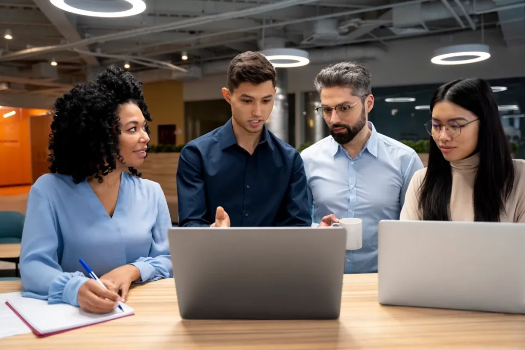 Quatro profissionais de negócios reunidos em um ambiente de escritório, analisando dados e discutindo ideias em frente a laptops.