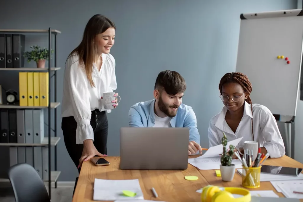 Três colegas de trabalho reunidos em uma mesa no escritório, analisando documentos e trabalhando em equipe com um laptop.