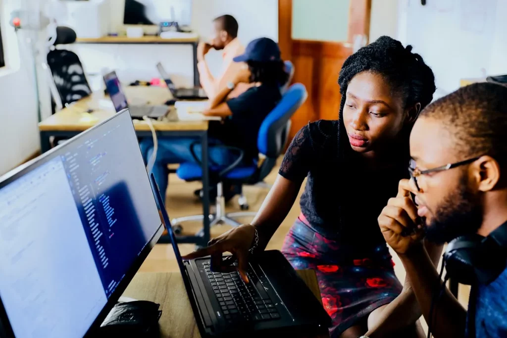 Dois colegas de trabalho colaborando em frente a um computador em um escritório moderno, simbolizando trabalho em equipe e troca de conhecimento.