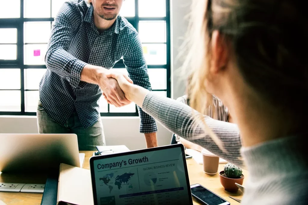 Duas pessoas apertando as mãos em um ambiente corporativo, com um laptop exibindo dados de crescimento da empresa em primeiro plano.