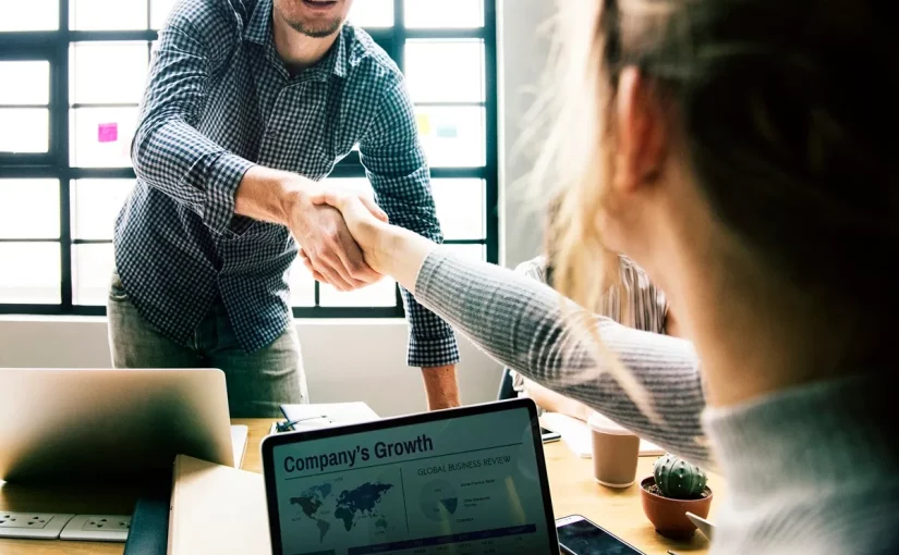 Duas pessoas apertando as mãos em um ambiente corporativo, com um laptop exibindo dados de crescimento da empresa em primeiro plano.