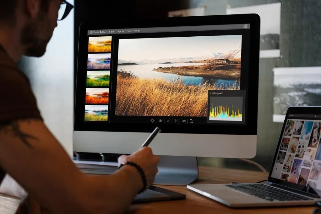 Pessoa usando uma caneta digital para editar uma foto em um monitor com ferramentas de edição e histogramas, com um laptop ao lado.