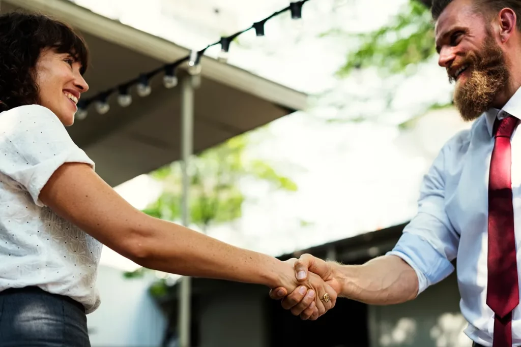 Duas pessoas sorridentes apertando as mãos em um ambiente externo, simbolizando uma parceria ou acordo profissional.