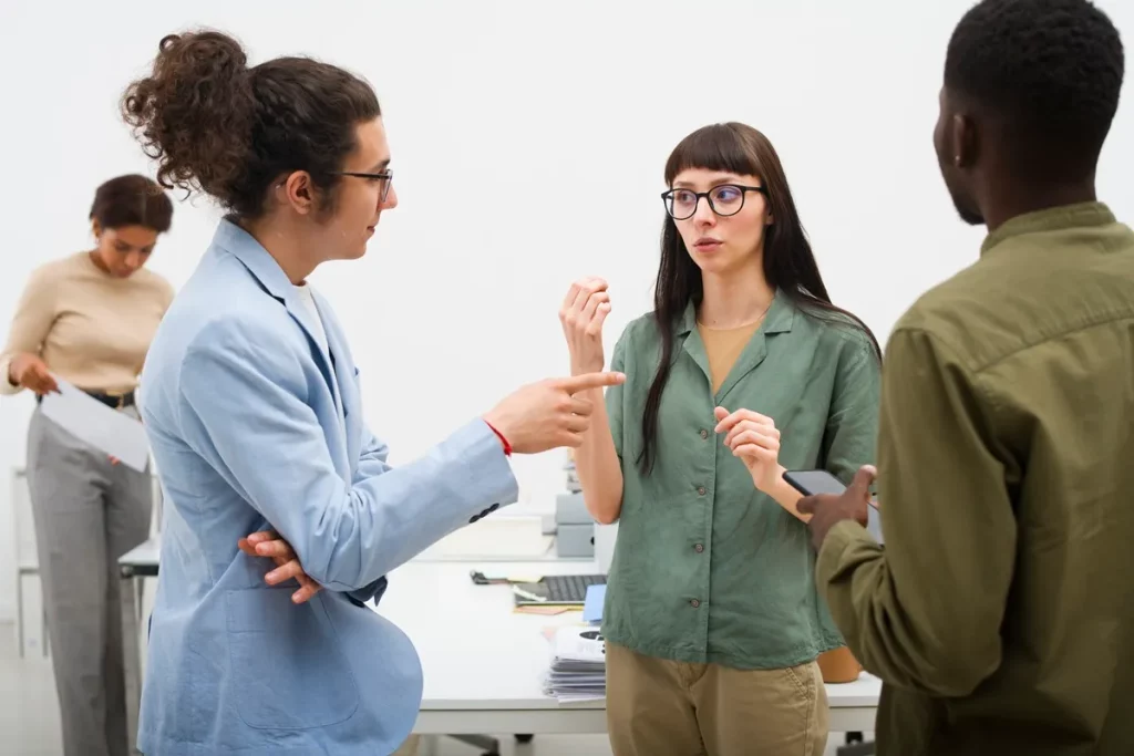 Três pessoas em um ambiente corporativo discutindo ideias, enquanto uma quarta pessoa trabalha ao fundo. A interação demonstra o uso da linguagem corporal em conversas profissionais.