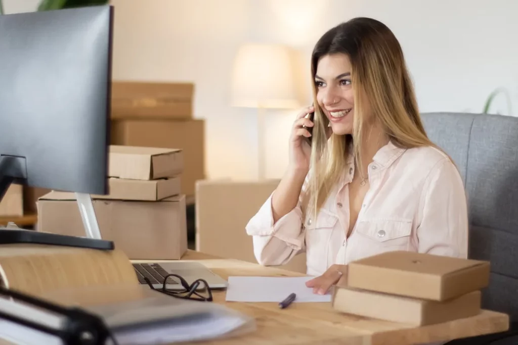 Mulher sorridente ao telefone em escritório com caixas de encomendas e computador, representando atendimento ao cliente e administração de loja virtual.