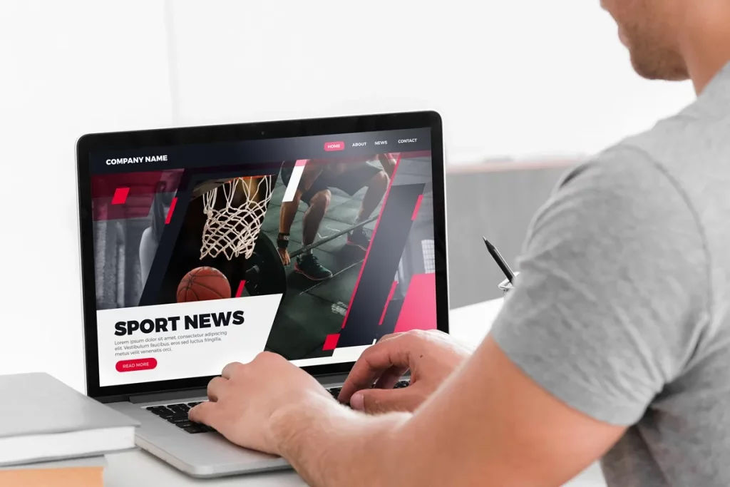 Pessoa acessando uma página de notícias esportivas em laptop com imagens de basquete e design moderno em vermelho e preto