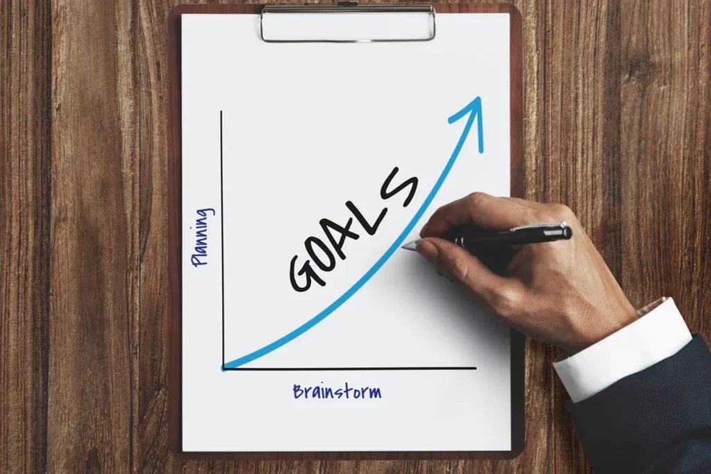Mão desenhando uma linha crescente em um gráfico com a palavra "Goals" em destaque, representando o alcance de metas através de planejamento e brainstorming.