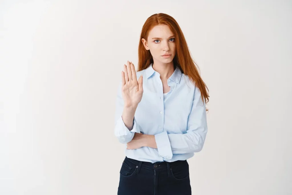 Mulher ruiva de expressão séria com a mão levantada em sinal de "pare", vestindo camisa azul clara e calça escura. Fundo branco minimalista.