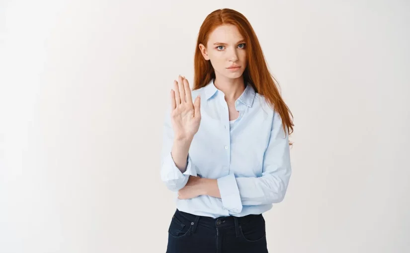 Mulher ruiva de expressão séria com a mão levantada em sinal de "pare", vestindo camisa azul clara e calça escura. Fundo branco minimalista.