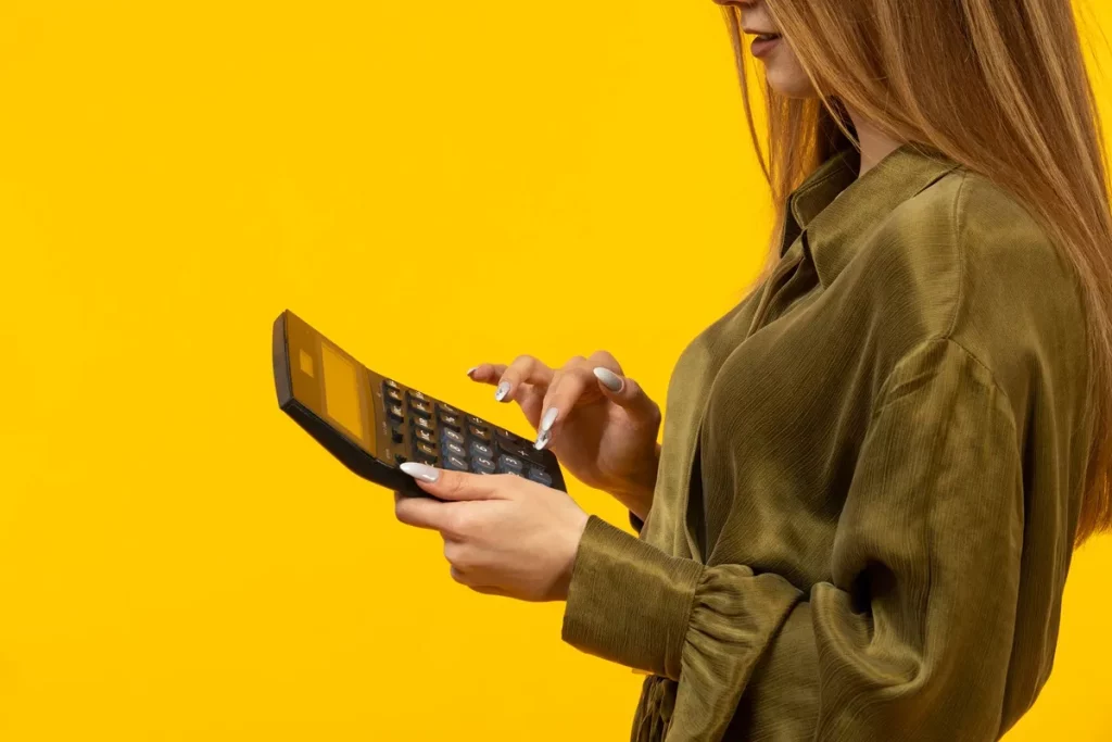 Mulher com camisa verde usando uma calculadora sobre fundo amarelo, simbolizando controle financeiro e análise de despesas