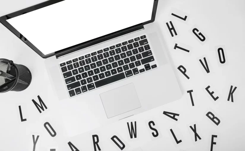 Laptop prateado sobre uma mesa branca com várias letras espalhadas ao redor, representando a escolha de palavras-chave para SEO.